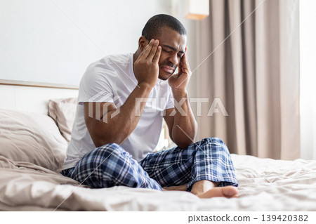 African american man sitting on bed holding temples with pain in bedroom African american man sitting on bed holding temples with pain in bedroom 139420382