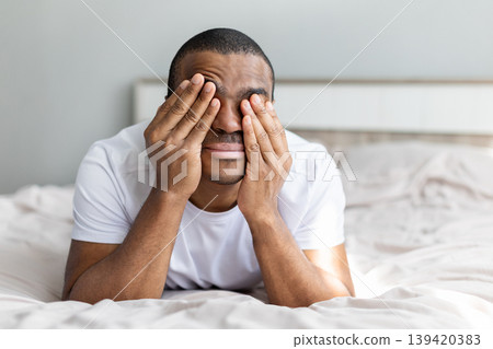 African american man lying on bed covering face with hands, expressing stress 139420383