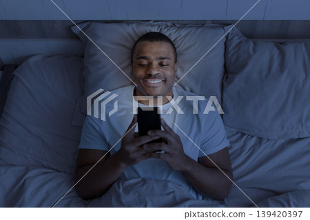 Smiling African american man using smartphone in bed at night in darkness Smiling African american man using smartphone in bed at night in darkness 139420397