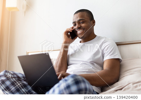 African american man talking on smartphone while using laptop on bed African american man talking on smartphone while using laptop on bed 139420400