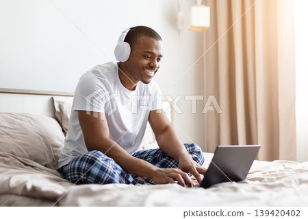 African american man sitting on bed typing on laptop in bedroom 139420402