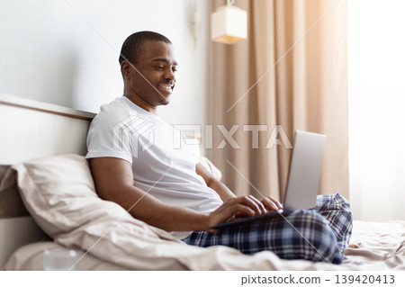 Happy African American man smiling while using laptop in bright morning bedroom Happy African American man smiling while using laptop in bright morning bedroom 139420413