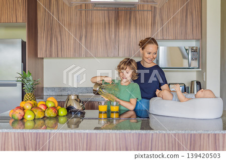 Mother and son preparing smoothie in kitchen while baby lies in cozy lounger on table watching. Family cooking together, healthy lifestyle, parenting, sibling bonding and everyday life concept 139420503
