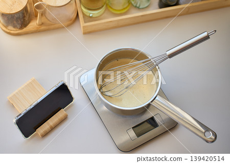 Saucepan with whisk and creamy mixture on kitchen scale near smartphone 139420514