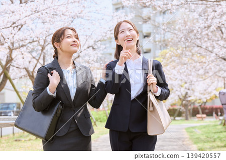 Two young businesswomen, dressed in suits and likely new employees, walk under a row of cherry trees, creating a spring commute scene. 139420557