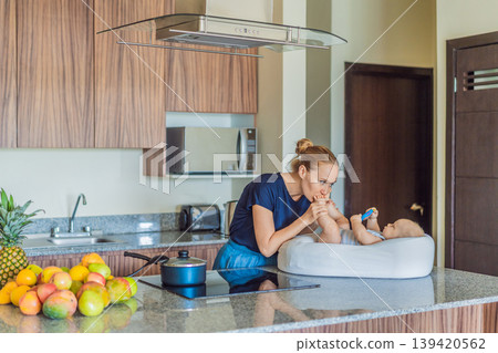 Mother talking to baby lying in cozy lounger on kitchen table while cooking at home. Caring mom interacting with infant during daily routine. Parenting, multitasking and family life concept 139420562