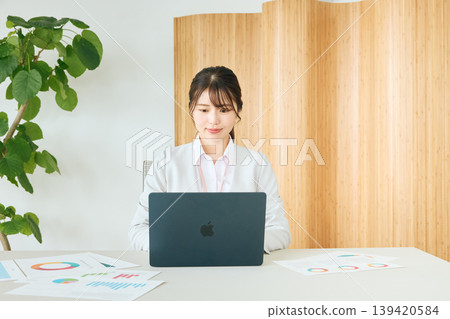 一位年輕的女商務人士正在辦公室使用筆記型電腦查看文件;一位身穿白色西裝的公司員工。 一位年輕的女商務人士正在辦公室使用筆記型電腦查看文件;一位身穿白色西裝的公司員工。 139420584