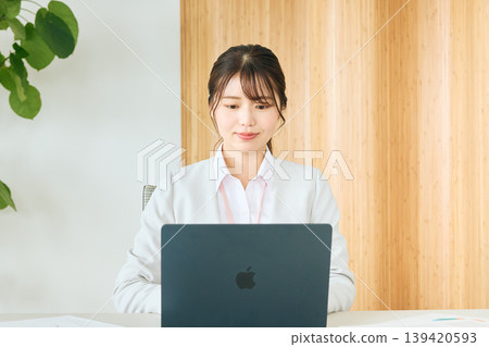 A young Japanese female business person working on a laptop in the office; a company employee in a white suit. 139420593