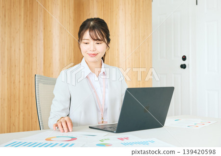 A young Japanese female businessperson working in an office using a laptop and documents; a company employee in a suit. A young Japanese female businessperson working in an office using a laptop and documents; a company employee in a suit. 139420598