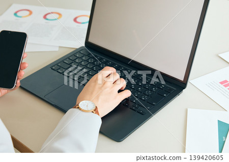 A businesswoman working at her desk using a laptop and smartphone; her hands, documents, and graphs are visible. 139420605