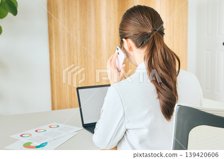 A businesswoman's back view as she uses her laptop while talking on her smartphone in the office. A businesswoman's back view as she uses her laptop while talking on her smartphone in the office. 139420606