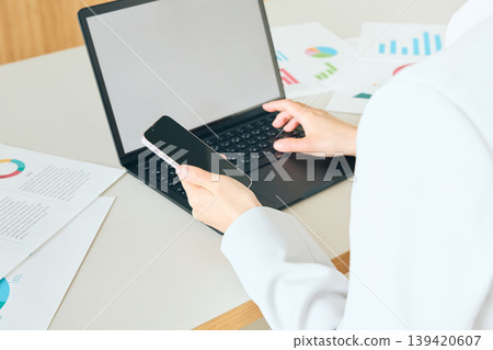 A businesswoman operating a laptop and smartphone; a female office worker analyzing data at her desk. 139420607