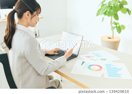 A young female business professional uses a laptop and documents in the office to analyze data, checking graphs and charts. 139420610