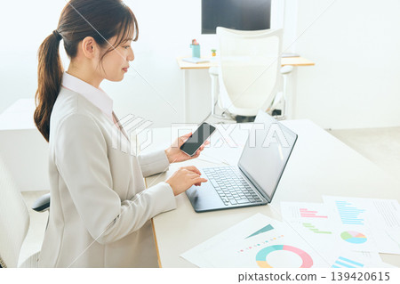 A young female business professional is reviewing data analysis documents using her smartphone and laptop in the office. 139420615