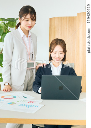Two young female business professionals are smiling and discussing a presentation in an office. Two young female business professionals are smiling and discussing a presentation in an office. 139420619