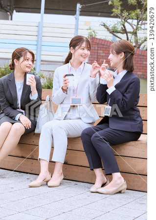 三位年輕的職業婦女(上班族)在午休時間坐在戶外長椅上聊天,並查看她們的智慧型手機。 三位年輕的職業婦女(上班族)在午休時間坐在戶外長椅上聊天,並查看她們的智慧型手機。 139420679