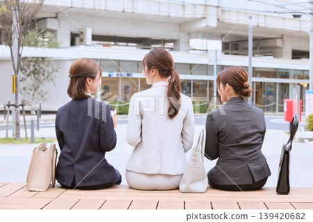 The backs of three young women in suits, business professionals, sitting on a bench and chatting in the square in front of the station. 139420682