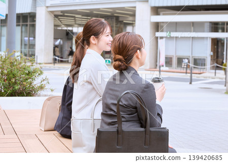 Two young businesswomen in suits are sitting on a bench in front of the station, chatting and taking a break with coffee in hand. Two young businesswomen in suits are sitting on a bench in front of the station, chatting and taking a break with coffee in hand. 139420685