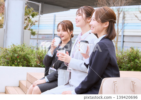 Three young female business professionals and a new employee in a suit are taking a break on an outdoor bench, drinking coffee. Three young female business professionals and a new employee in a suit are taking a break on an outdoor bench, drinking coffee. 139420696