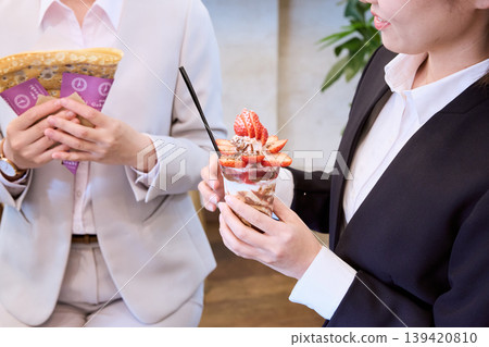 Two women in suits holding sweets; businesswomen enjoying a strawberry parfait and crepes during their break. Two women in suits holding sweets; businesswomen enjoying a strawberry parfait and crepes during their break. 139420810