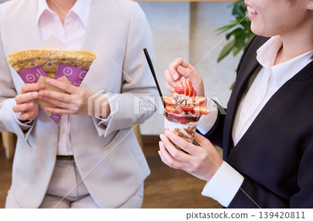 Two women in suits, holding crepes and strawberry parfaits, are seen having lunch at a cafe – businesswomen. 139420811