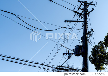 Silhouette of an electrical utility pole with a transformer and complex network of power lines against a clear blue sky. Industrial infrastructure and urban energy distribution concept. Silhouette of an electrical utility pole with a transformer and complex network of power lines against a clear blue sky. Industrial infrastructure and urban energy distribution concept. 139422292