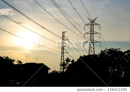 Silhouette of high voltage electricity transmission towers and power lines against a glowing sunset. Industrial energy infrastructure landscape with trees and residential building in foreground. Silhouette of high voltage electricity transmission towers and power lines against a glowing sunset. Industrial energy infrastructure landscape with trees and residential building in foreground. 139422300