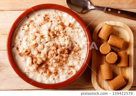 A brown ceramic bowl filled with creamy white corn porridge and cinnamon powder, served with peanut brittle and a spoon on a rustic wooden table A brown ceramic bowl filled with creamy white corn porridge and cinnamon powder, served with peanut brittle and a spoon on a rustic wooden table 139422578