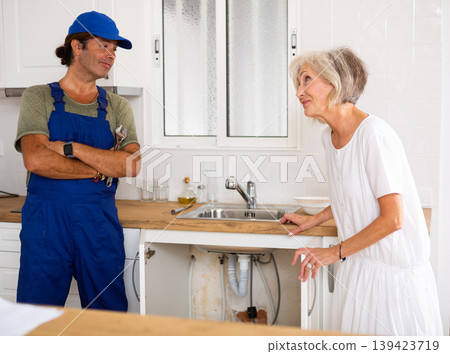 Elderly woman thanking professional plumber for work in kitchen Elderly woman thanking professional plumber for work in kitchen 139423719