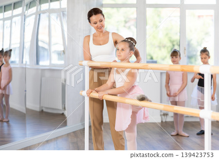 Girl ballet students prepare for classes, near ballet bare stretch their leg under teacher guidance. 139423753