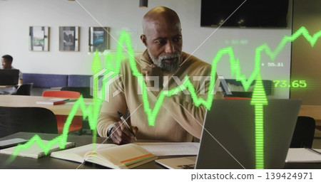 Senior exec in tan sweater writing with pen reviewing laptop at coworking space, with chart overlay Senior exec in tan sweater writing with pen reviewing laptop at coworking space, with chart overlay 139424971