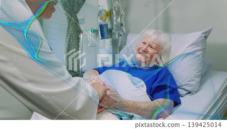 Patient lying on hospital bed in blue gown smiling at nurse holding hand, beside IV drip 139425014