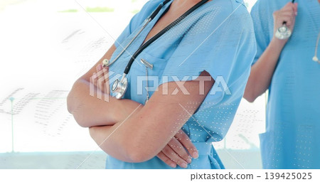 Standing nurse in scrubs crossing arms, colleague holding stethoscope in hospital hallway, sunlight Standing nurse in scrubs crossing arms, colleague holding stethoscope in hospital hallway, sunlight 139425025