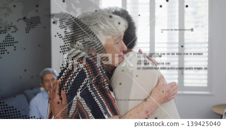 Hugging two senior women wearing striped blouse and knit sweater in living room, with sofa shutters Hugging two senior women wearing striped blouse and knit sweater in living room, with sofa shutters 139425040