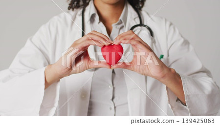 Holding red heart, doctor wearing white coat and stethoscope against gray wall, heart health Holding red heart, doctor wearing white coat and stethoscope against gray wall, heart health 139425063