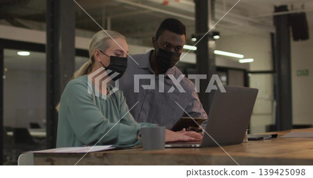 Collaborating masked colleagues examining smartphone typing at office desk with laptop coffee mug Collaborating masked colleagues examining smartphone typing at office desk with laptop coffee mug 139425098