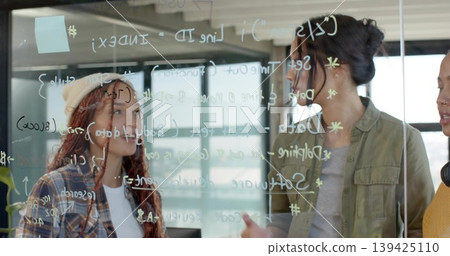 Women in varied attire collaborating on code behind glass board in office, with pastel sticky note 139425110