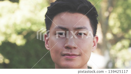 Showing adult Asian man wearing dark shirt in park under sunlight, showcasing blurred green foliage Showing adult Asian man wearing dark shirt in park under sunlight, showcasing blurred green foliage 139425117