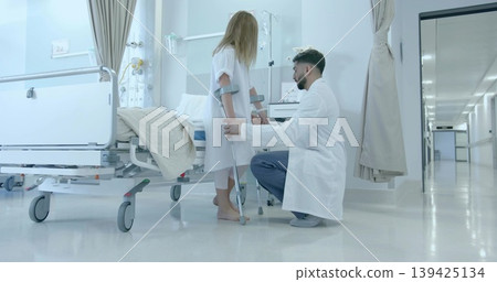 Kneeling doctor helping child in gown using crutches in ward, with bed, IV pole, cart, curtains Kneeling doctor helping child in gown using crutches in ward, with bed, IV pole, cart, curtains 139425134