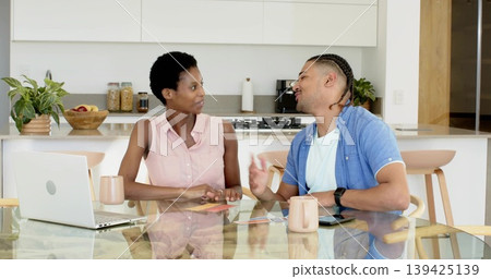 Discussing couple reviewing laptop and checking phone at modern kitchen table, with coffee mugs Discussing couple reviewing laptop and checking phone at modern kitchen table, with coffee mugs 139425139