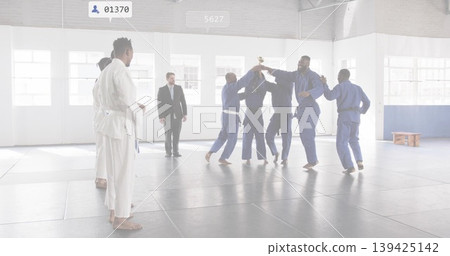 Six martial artists in blue uniforms lifting gold trophy in dojo, instructor in white gi observing 139425142