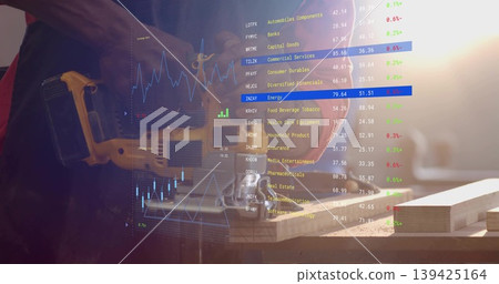 Woodworker cutting plank with yellow jigsaw saw on workshop bench, stock market overlay, copy space 139425164