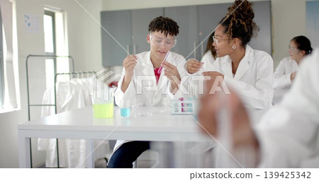 Using dropper and test tube, lab partners wearing labcoats goggles at school lab with neon beakers Using dropper and test tube, lab partners wearing labcoats goggles at school lab with neon beakers 139425342