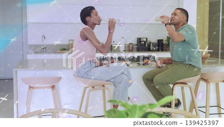 Two friends drinking glass water bottles in home kitchen with island, with light wood bar stools Two friends drinking glass water bottles in home kitchen with island, with light wood bar stools 139425397
