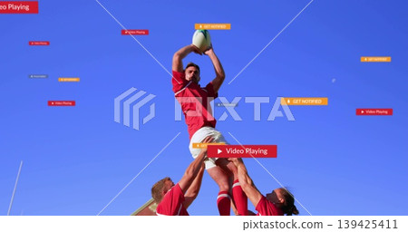 Rugby player in red jersey catching white-green ball as teammates lifting him on pitch, goal post 139425411
