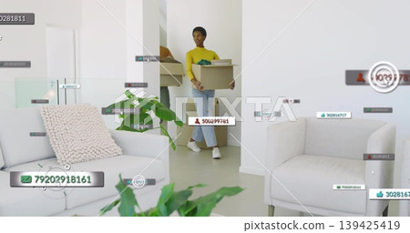 Walking mid-adult woman carrying box through modern living area, with digital overlay icons Walking mid-adult woman carrying box through modern living area, with digital overlay icons 139425419
