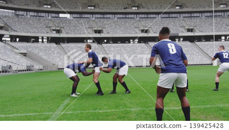 Forming seven male athletes practicing scrum in empty stadium, with blue jerseys and white shorts 139425428