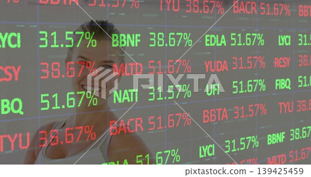 Smiling woman standing behind transparent display at trading floor, displaying tickers in red green Smiling woman standing behind transparent display at trading floor, displaying tickers in red green 139425459