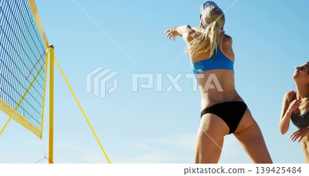 Leaping female athlete striking blue-and-white volleyball at beach court, with yellow net 139425484