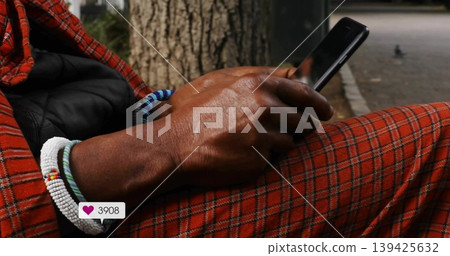 Scrolling man wearing plaid pants beside tree trunk, with heart icon overlay on phone and bracelets 139425632